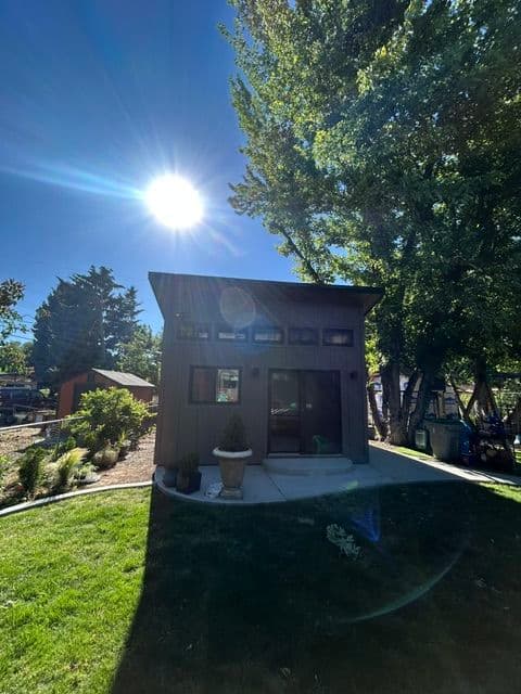 Modern small house with large windows under clear blue sky, surrounded by greenery.