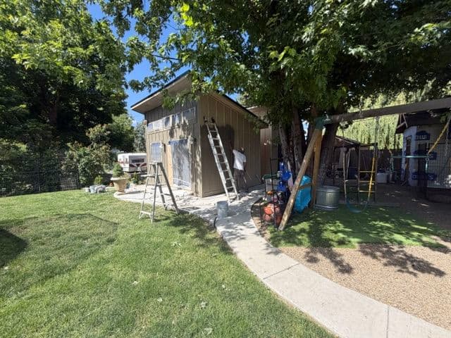 Person painting a shed in a backyard with a ladder and playground equipment nearby.