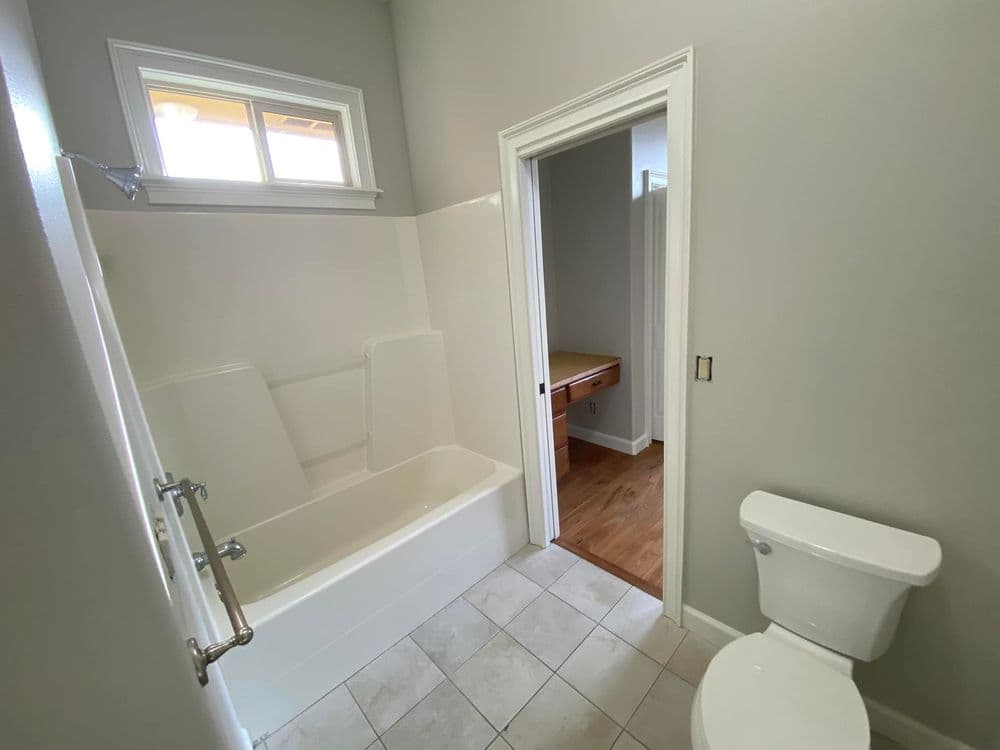 Modern bathroom with beige tile flooring, bathtub, toilet, and adjoining room.