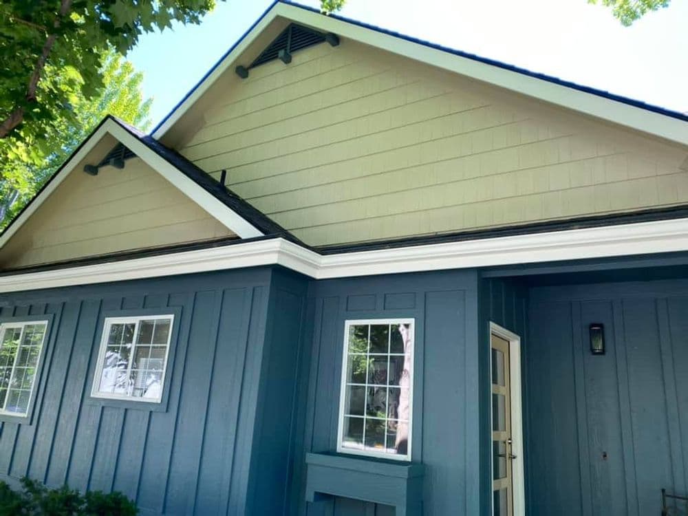 Blue and beige modern house with a gabled roof and large windows, surrounded by trees.